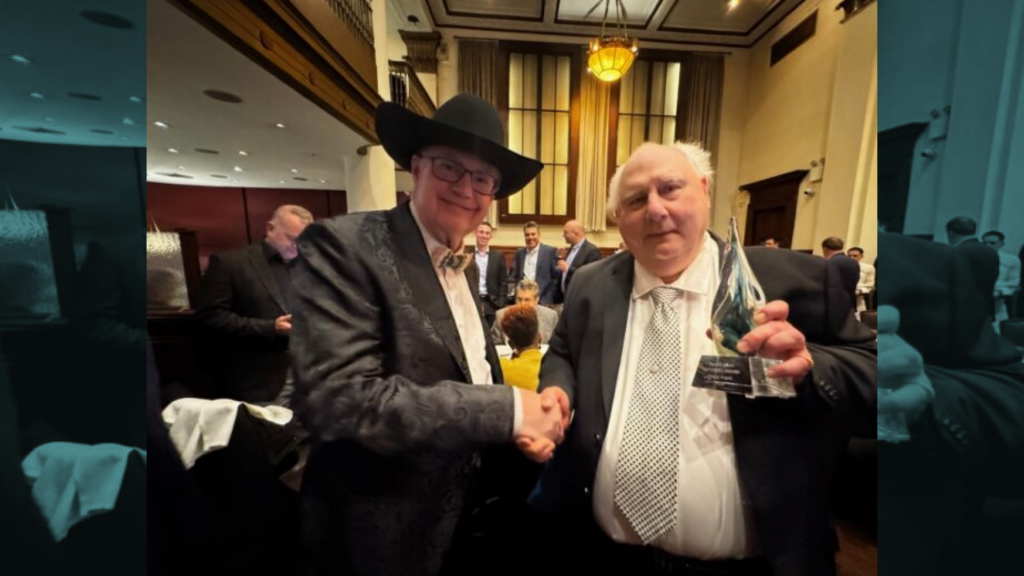 Two men in formal suits shake hands at an awards ceremony; one wears a black cowboy hat while the other holds a glass award plaque.